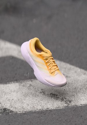 Scarpa da ginnastica gialla e bianca con tomaia texturizzata, tre strisce bianche e suola ammortizzata, mostrata in aria contro una superficie grigia.