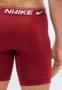 Κοντινό πλάνο ενός ατόμου που φοράει κόκκινα ανδρικά εσώρουχα Nike με ορατό λογότυπο στη μέση και μέρος του χεριού τους να ακουμπάει δίπλα τους.