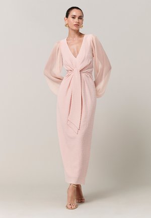 Ženska, oblečena v svetlo rožnato midi obleko z teksturo, prosojnimi dolgimi nabranimi rokavi, globokim V-izrezom, pasnim trakom, zlatimi uhani in zlatimi sandali s peto.