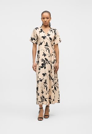 Kvinne med flettehår iført en beige omslagskjole med svart blomstertrykk og svarte sko med ankelstropp, stående foran en ensfarget bakgrunn.