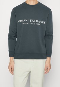 Sweatshirt verde scuro con scollatura tonda, maniche lunghe e polsini a coste; presenta la scritta bianca "Armani Exchange Milano/New York" sul davanti.