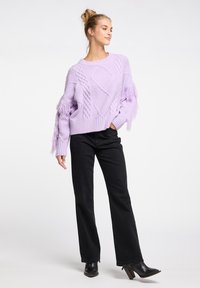 Lila Strickpullover mit strukturierten Kabelmustern und Fransendetails an den Ärmeln, kombiniert mit schwarzen weit geschnittenen Jeans und knöchelhohen Stiefeln mit Absatz.