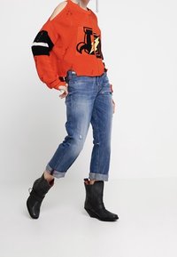 Pull en tricot rouge avec un motif d'éclair, à l'aspect usé ; associé à un jean en denim déchiré bleu clair et des bottines noires.