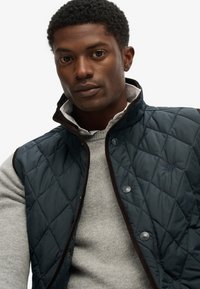 Quiltet mørkeblå vest med høj krave, brune accenter og knaplukninger over en grå sweater. Har et syet diamantmønster og logo-detalje.