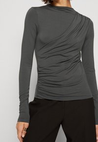 Kvinna som bär en åtsittande långärmad topp i kolgrått med draperade detaljer och svarta byxor, med handen avslappnad vid sidan och en guldring.