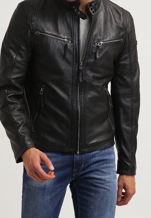 Mężczyzna w czarnej skórzanej kurtce z kieszeniami na zamek i ciemnoniebieskich dżinsach, stojący na jasnym, jednolitym tle.