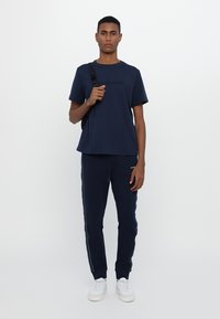 Marineblå t-skjorte med teksturert logo, kombinert med matchende joggebukser med hvite sidelister. Modellen bruker hvite sneakers og en svart ryggsekk.