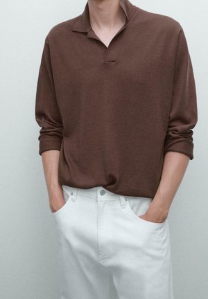 Personne portant une chemise marron à manches longues avec un col et un pantalon blanc, les mains posées avec décontraction dans les poches, debout devant un fond uni.