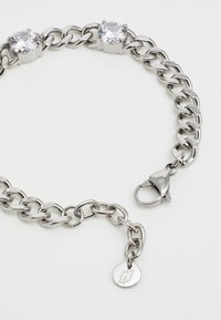 Bracciale in catena di metallo argentato con due gemme trasparenti, chiusura a gancio e un piccolo tag con logo. Finitura liscia con anelli ovali.
