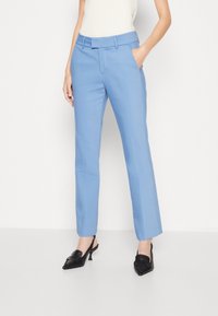 Svetle modré kalhoty na míru se rovný fit, přední kapsy, poutka na opasek a hladká textilie. Nosené s černými lodičkami.