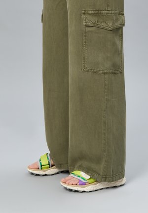 Pantaloni verde oliva a gamba larga che coprono sandali aperti multicolore con cinturini verdi, viola e beige su uno sfondo neutro.