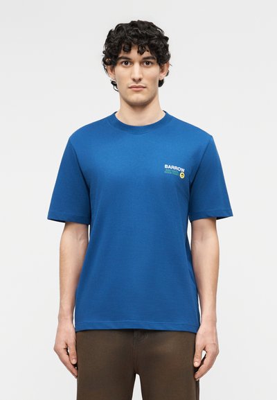 Νεαρός άνδρας με σγουρά σκούρα μαλλιά, φοράει μπλε μπλουζάκι Barrow και καφέ παντελόνι, στέκεται μπροστά σε ένα απλό φωτεινό φόντο.