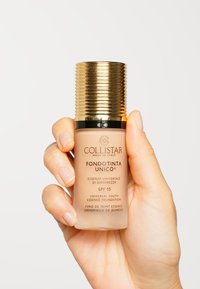Flacon de fond de teint en forme cylindrique, en verre beige avec des accents dorés, capuchon doré strié, étiqueté avec les détails du produit et SPF 15.