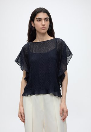 Jeune femme portant un chemisier court à manches courtes en tissu transparent bleu marine texturé, par-dessus un caraco, assorti à un pantalon blanc taille haute sur fond blanc.