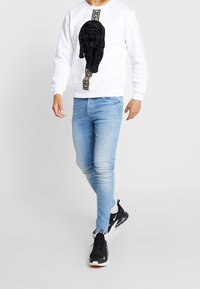 Muž jde v bílém mikině s grafickým motivem černého pantera, světle modrých skinny džínách a černých teniskách Nike s bílou podrážkou.