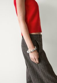 Débardeur rouge en maille, pantalon gris à fines rayures, et un bracelet chunky argenté, mettant en valeur des textures métalliques lisses et des formes courbes.