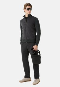 Tmavá bunda na zip s prošívanou přední částí, šedé rukávy, černé kalhoty, béžové tenisky a malá černá taška. Hladké a texturované materiály.