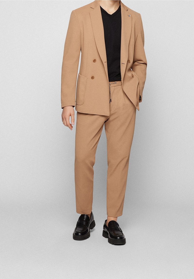 Homme portant un blazer croisé couleur beige et un pantalon assorti, avec une chemise noire à col en V et des mocassins en cuir noir.