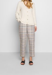 Cremefarbener Cardigan mit Knöpfen und zwei Fronttaschen, kombiniert mit karierten Hosen in Grau, Braun und Elfenbein, getragen mit schwarzen Pantoletten.