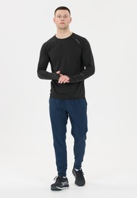 Langärmliges schwarzes Sportoberteil aus strukturiertem Stoff, kombiniert mit navyblauen, taillierten Hosen, ergänzt durch schwarze Laufschuhe.