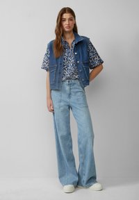 Denim vest met een kraag en knopen aan de voorkant, gecombineerd met een blauwe bloemetjesoverhemd met korte mouwen en lichtblauwe wijde jeans. Witte sneakers.