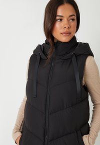 Frau trägt eine schwarze, ärmellose, gesteppte Weste mit Kapuze und Reißverschluss, beige langärmliges Oberteil, Hände in den Taschen, schaut zur Seite.