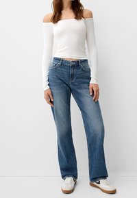 Mujer vestida con una blusa blanca de manga larga y sin hombros, jeans rectos de talle alto azules y zapatillas blancas, contra un fondo liso.
