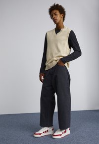 Beige knit vest with a V-neck over a black long-sleeve shirt, paired with loose gray trousers and white sneakers with red stripes.