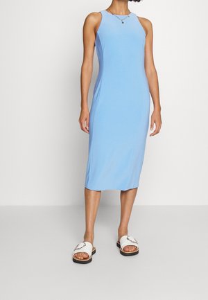 Woman wearing a sleeveless light blue midi dress and white slide sandals standing against a plain background.