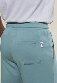 Nahaufnahme der tealfarbenen Sweatpants mit Gesäßtasche und Kangaroos-Marke, getragen mit einem weißen Shirt, das den elastischen Bund und die Nähdetails zeigt.