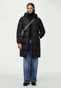 Melna izšūta gara virsjaka ar rāvējslēdzēju priekšpusē un kapuci. Valkāta pār melnu augstu apkaklīti, kopā ar platajiem zilajiem džinsiem un leopardprinta somu.