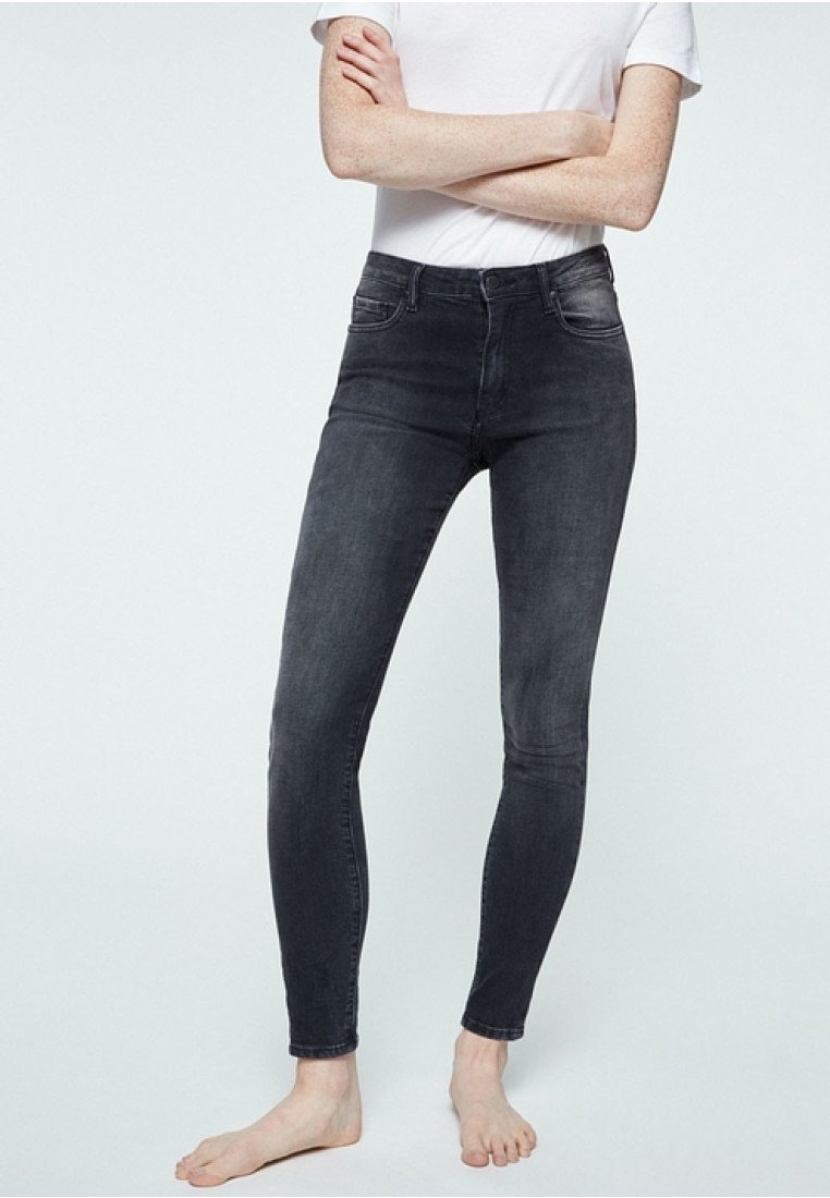 Personne pieds nus portant un jean noir ajusté et une chemise blanche à manches courtes, les bras croisés, debout devant un fond clair uni.