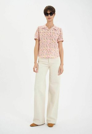 Blouse rose à manches courtes avec un col, ornée de fleurs, associée à un pantalon large crème et des chaussures marron, portée par un mannequin avec des lunettes de soleil.