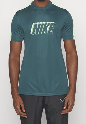 Mężczyzna w ciemnozielonym koszulce Nike z krótkim rękawem z jasnooliwkowymi akcentami oraz czarnych spodniach z białym logo Nike.