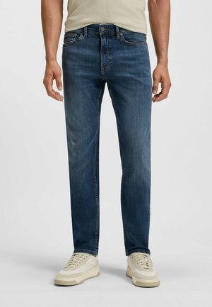 Jean en denim bleu foncé avec une coupe droite. Présente un design classique, cinq poches et une légère décoloration. Associé à des baskets de couleur claire.