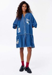 Džínové šaty s vrstveným dizajnom, predným gombíkom, krátkymi nadýchanými rukávmi a vyšívanými zvieracími motívmi. Dokončené kontrastným stehovaním.