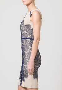 Beige šaty s námořnickou krajkovou vrstvou, přiléhavý střih, bez rukávů, délka po kolena, s horizontálním pasem a složitými krajkovými vzory.