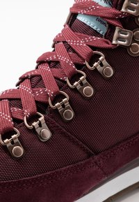 Bordeauxrode wandelschoen met een bovenwerk van gestructureerd mesh, metalen veterogen en contrasterende veters, voorzien van een gevoerde tong en een rubberen zool.