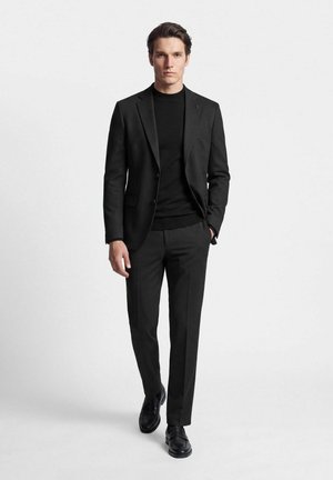Homme marchant vers l'avant portant un costume noir ajusté avec une chemise noire à col rond et des chaussures en cuir noir sur un fond blanc uni.