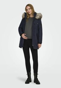 Námornícka parka s umelou kožušinou na kapucni, zapínanie na gombíky a zips, dve bočné vrecká a rebrované manžety. Nosené na šedom tričku a čiernych legínach, s čiernymi topánkami.