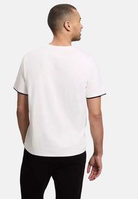 Biała koszulka z krótkim rękawem wykończona czarnym lamowanym wykończeniem, okrągłym dekoltem i luźnym krojem. Materiał wydaje się gładki i lekki.