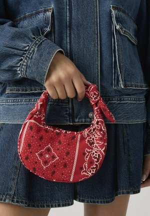 Mano sosteniendo un pequeño bolso de tela con estampado de bandana roja sobre una chaqueta y falda de mezclilla azul oscuro.