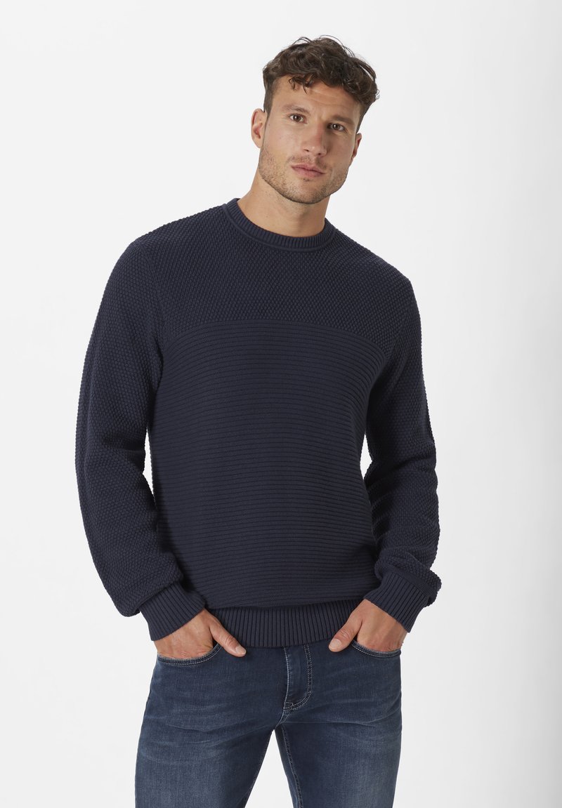 Marineblauer Strickpullover mit einem strukturierten Muster, ausgestattet mit einem gerippten Rundhalsausschnitt, langen Ärmeln und einem horizontal gestreiften Abschnitt, kombiniert mit Jeans.