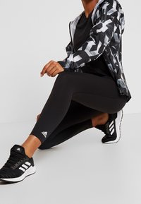 Leggings neri in tessuto testurizzato, abbinati a una giacca nera con motivo. Scarpe sportive nere con accenti bianchi e branding.