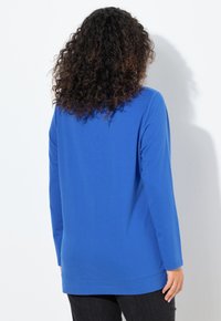 Modrý top s dlouhým rukávem z hladké tkaniny. Má volný střih a zakřivený lem, s jednobarevným designem bez dalších ozdob.