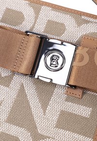 Ingrandimento di un tessuto beige intrecciato con grandi lettere bianche, una fascia color beige e una fibbia argentata lucida con un logo circolare in rilievo che presenta una "B".