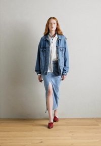 Denim jacket, white blouse, blue denim skirt with a slit, and red shoes. Model is standing on wooden floor against a light wall.