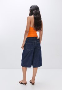 Oranje mouwloze top gecombineerd met donkerblauwe denim culottes. De culottes hebben een ontspannen pasvorm, zichtbare stiksels en achterzakken.