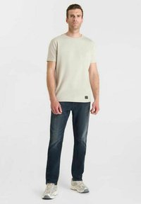 Uomo in piedi, con una maglietta a maniche corte beige chiaro, jeans blu scuro e sneakers bianche, su uno sfondo bianco uniforme.