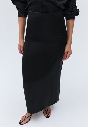 Femme portant une jupe midi noire ajustée avec un haut noir à manches longues et des sandales noires à brides, debout devant un fond uni.